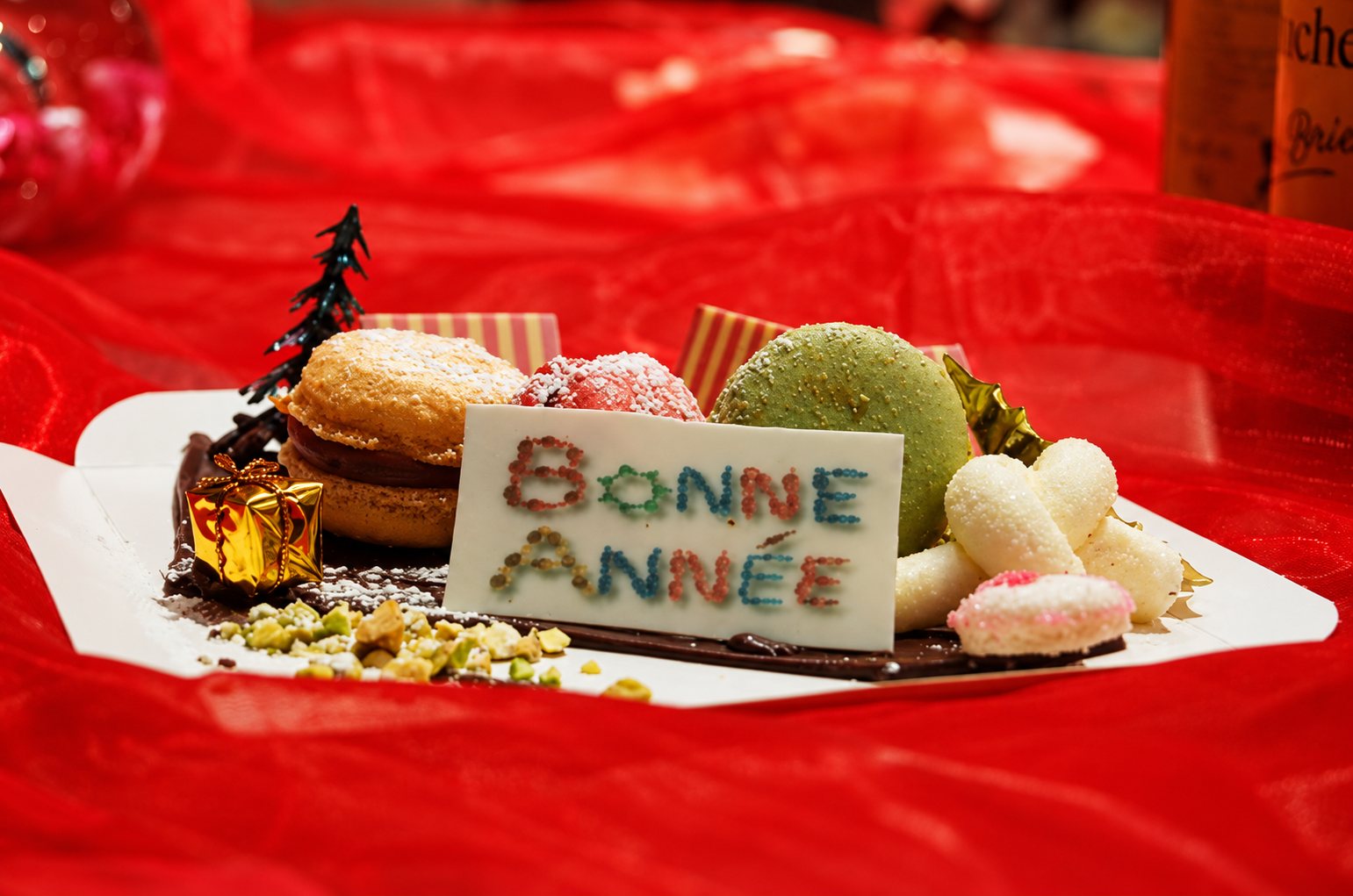 Une assiette de macarons et de petites douceurs décorée d'une plaquette en chocolat portant les mots Bonne Année, posée sur un voile rouge.
