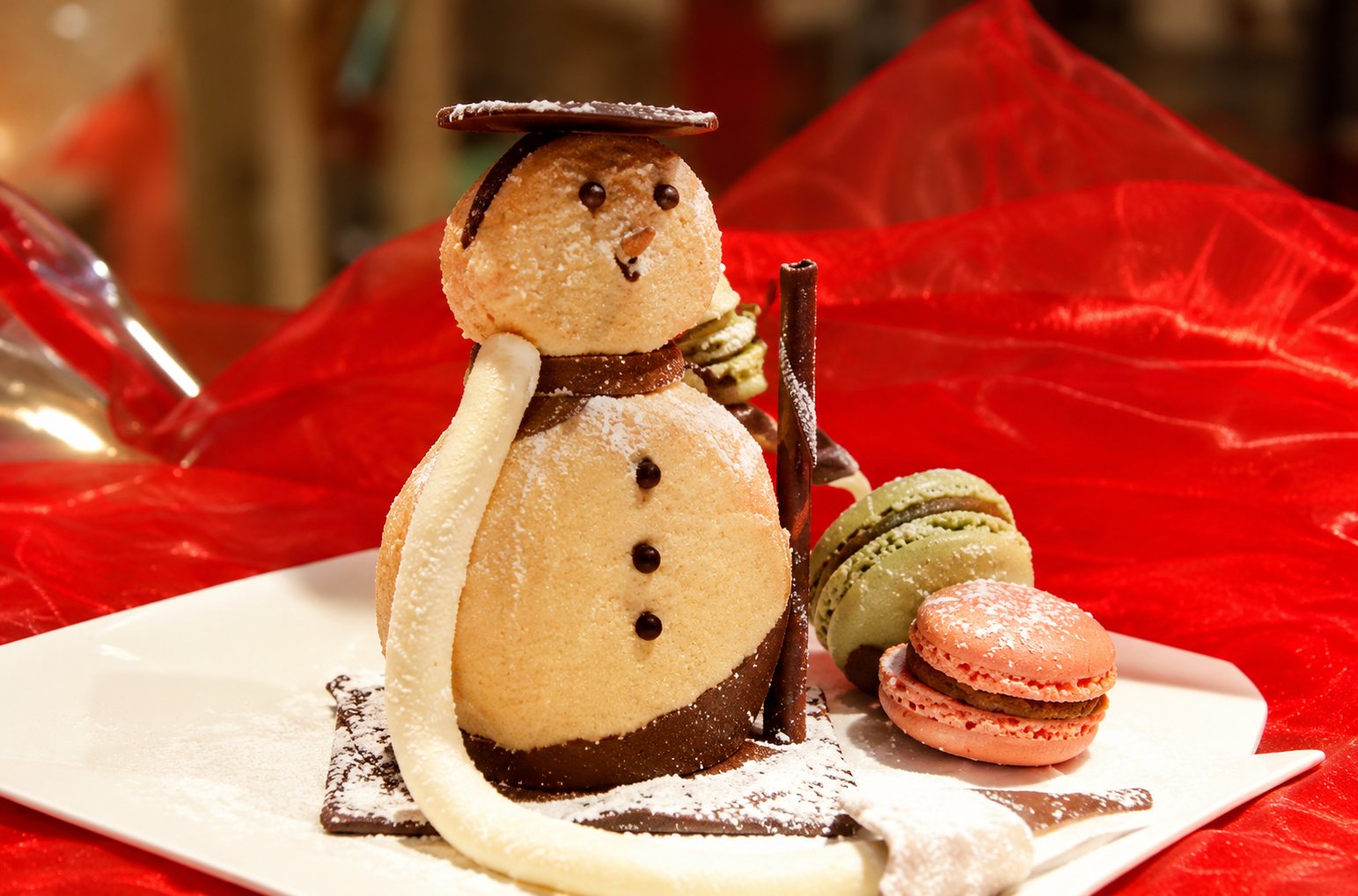 Un dessert en forme de bonhomme de neige en chocolat et blanc, posé sur une assiette blanche avec des macarons, sur un tissu rouge.
