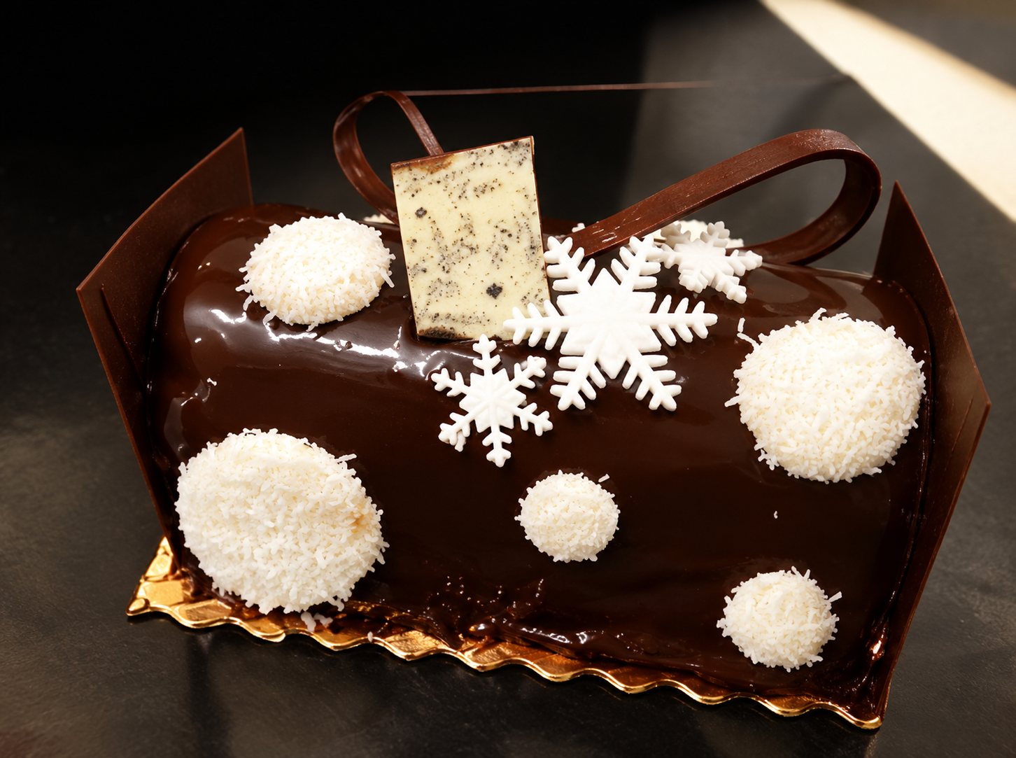 Une bûche de Noël glacée au chocolat, décorée de flocons de neige en sucre et de boules de noix de coco blanches, avec une petite plaquette dorée.