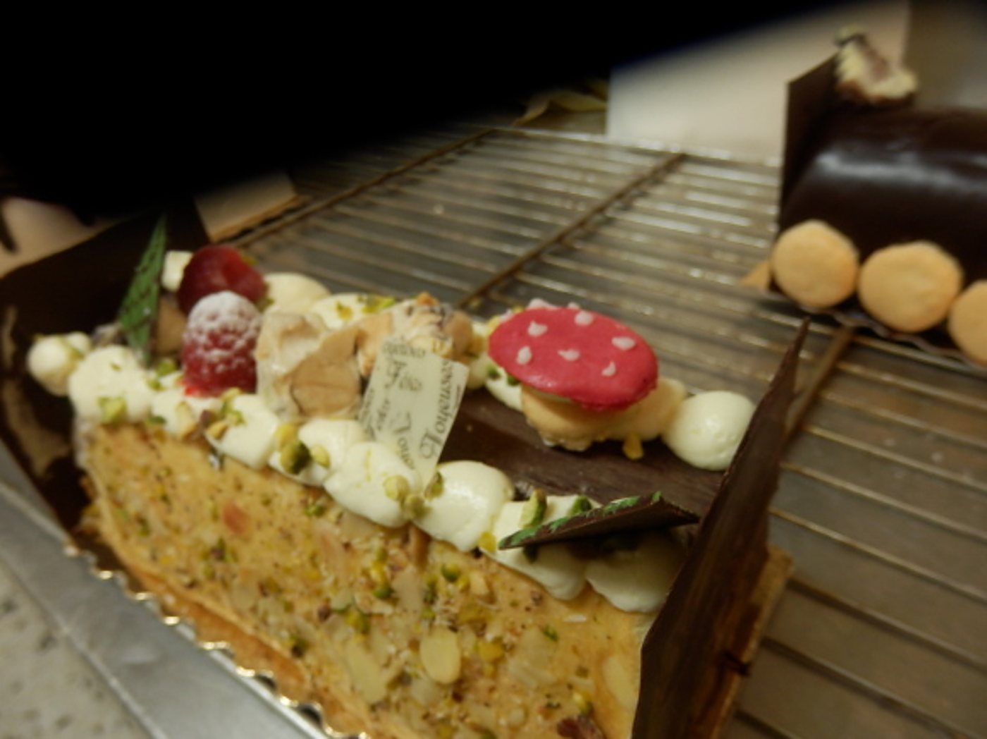 Une bûche de Noël pâtissière garnie de pistaches, de crème, de framboises et d'un champignon rouge, posée sur une grille de refroidissement.