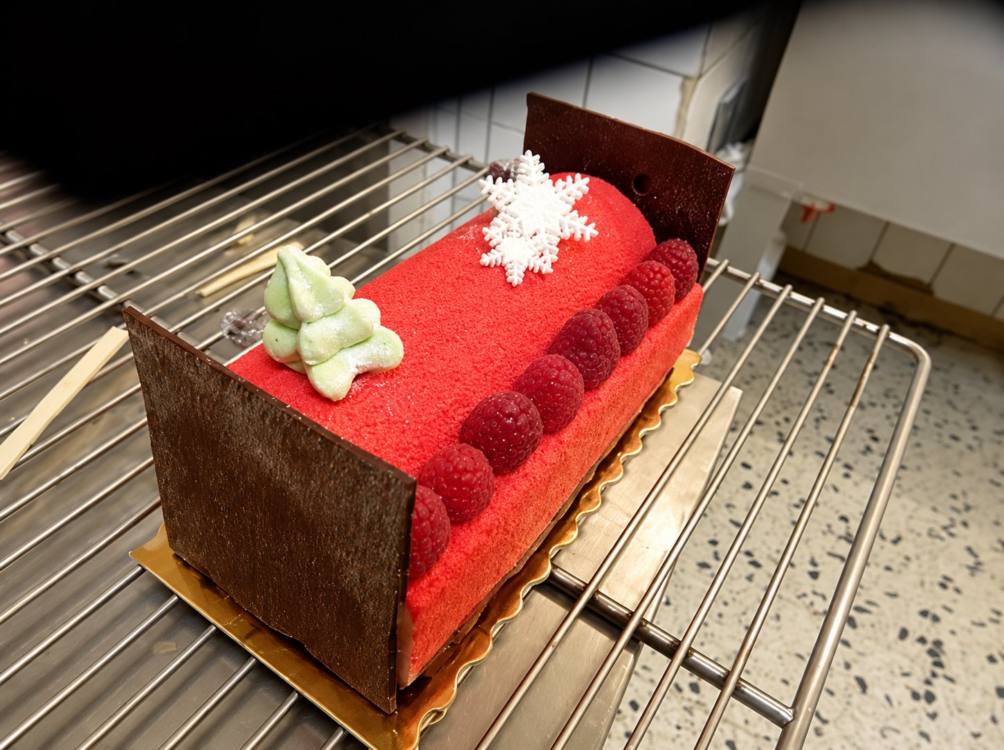 Une bûche de Noël glacée de rouge, ornée de framboises, d'un flocon blanc et d'un petit sapin vert, posée sur une grille de refroidissement.