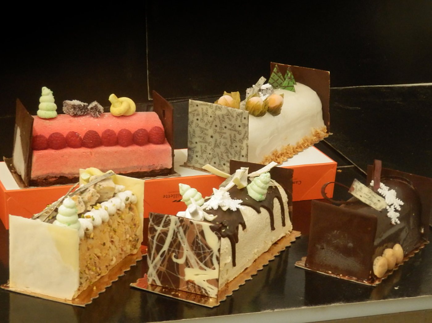 Cinq bûches de Noël glacées et chocolatées, décorées de framboises, de meringues vertes et de plaquettes en chocolat, posées sur des boîtes orange en vitrine.
