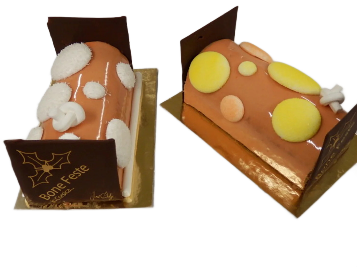 Deux bûches de Noël au glaçage orange décorées de pastilles blanches et jaunes, avec des plaques de chocolat portant l'inscription Bone Feste, posées sur du papier blanc.