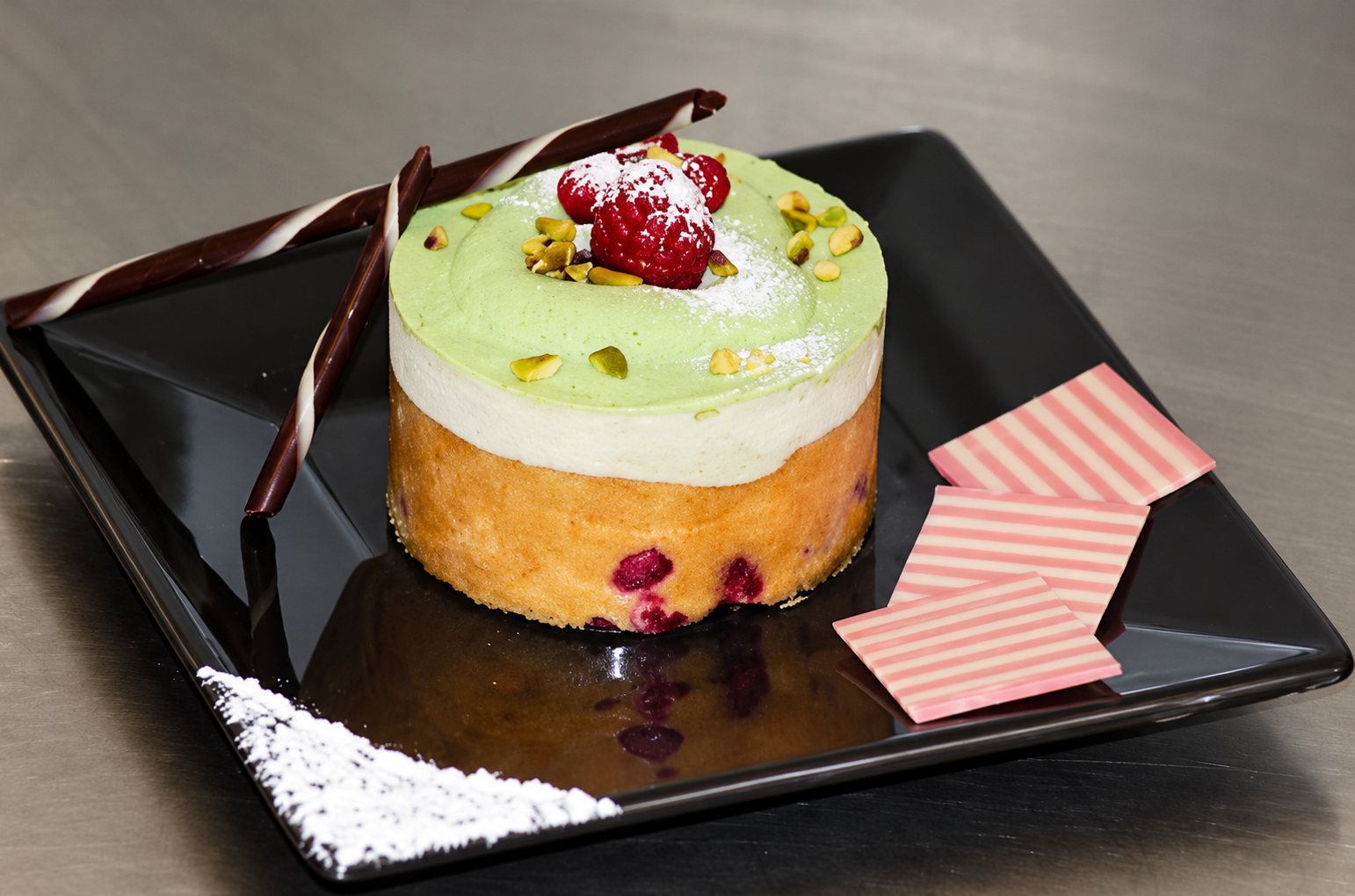 Un petit entremets rond surmonté d'une framboise et de pistaches, posé sur une assiette carrée noire avec des décors en chocolat rose et brun.