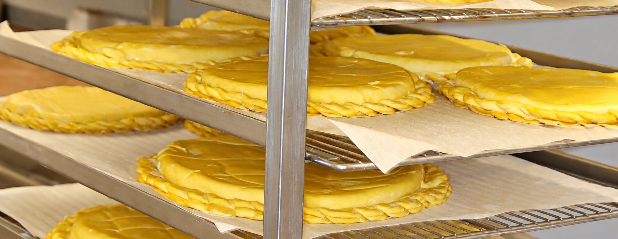Plusieurs galettes jaune doré au bord tressé reposent sur du papier sulfurisé, posées sur les étagères d'un chariot métallique de boulangerie.
