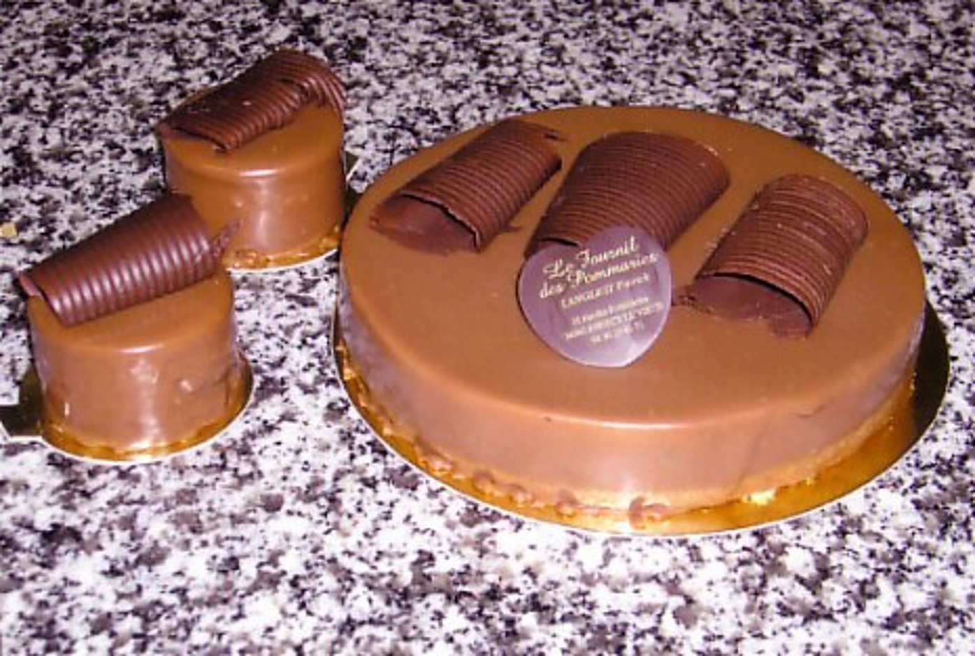 Un gâteau rond glacé de chocolat au lait décoré de copeaux de chocolat et d'un médaillon, posé près de deux petits entremets sur un plan en granit.
