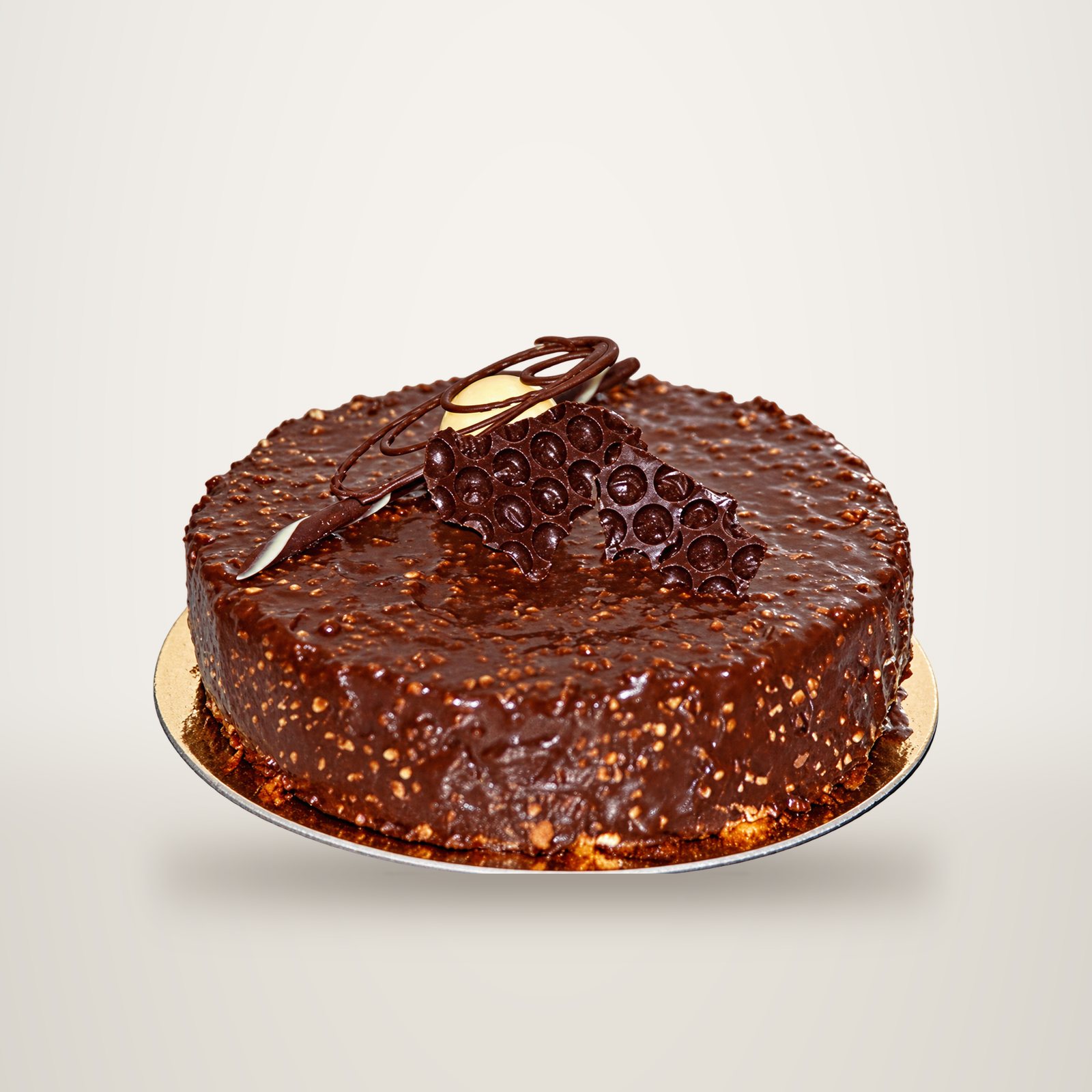 Un gâteau rond entièrement nappé de glaçage au chocolat et d'éclats de noisettes, décoré de ruban et de billes en chocolat, posé sur un support doré.