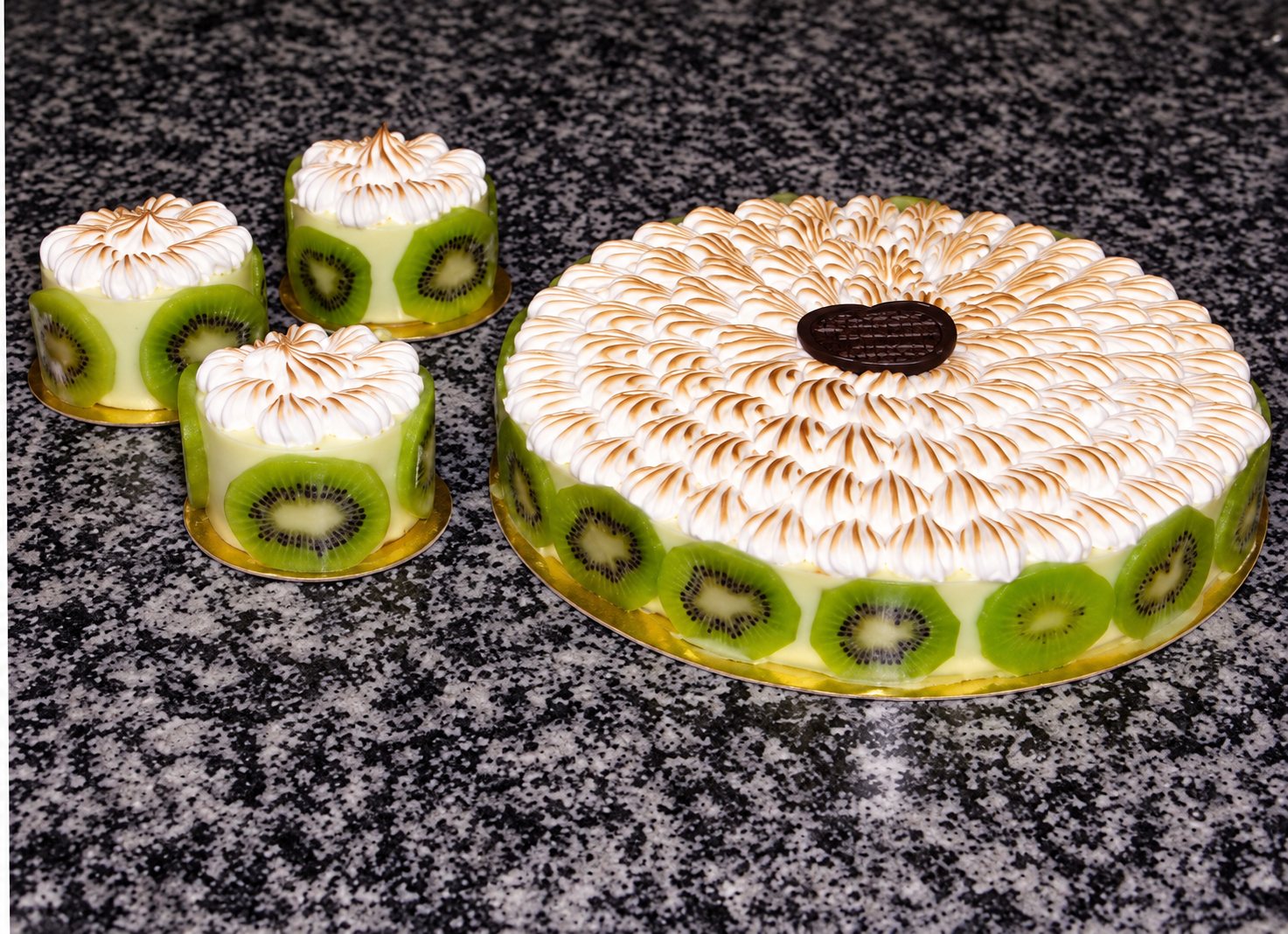 Un grand gâteau décoré de meringue et de tranches de kiwi posé à côté de trois petits entremets individuels sur un plan en granit.