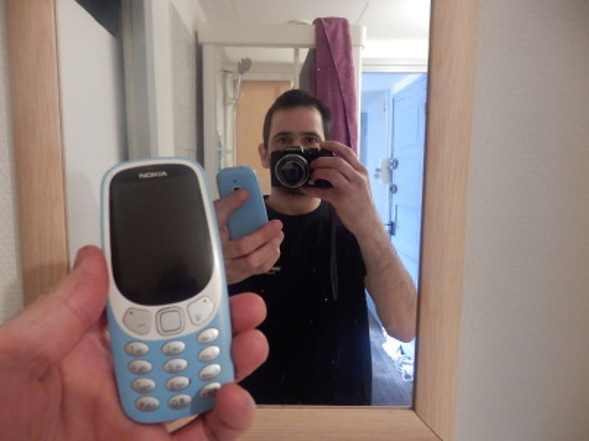 Un homme prend un selfie dans un miroir avec un appareil photo, tandis qu'une main au premier plan tient un téléphone Nokia bleu et qu'il en tient un autre.