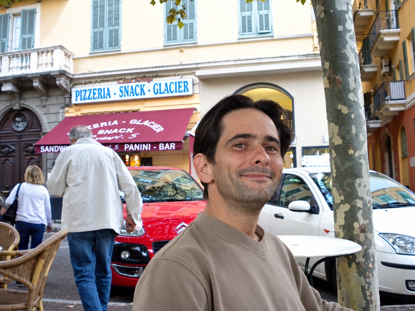 Un homme souriant est assis à une table en terrasse, devant la façade d'une pizzeria snack glacier avec un store rouge, tandis que des passants marchent derrière lui.