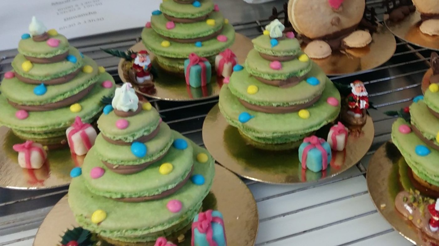 Plusieurs macarons verts empilés en forme de sapins de Noël, ornés de bonbons colorés, de petits cadeaux en pâte d'amandes et de Pères Noël, posés sur des disques dorés en vitrine.