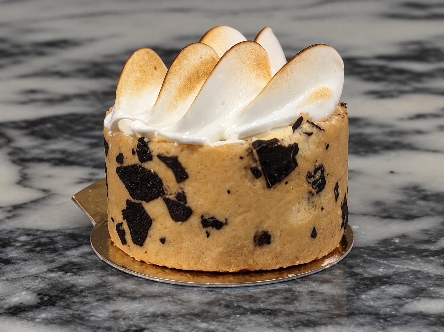 Un petit gâteau rond décoré de pétales de meringue dorés et de morceaux de chocolat, posé sur un comptoir en marbre gris.
