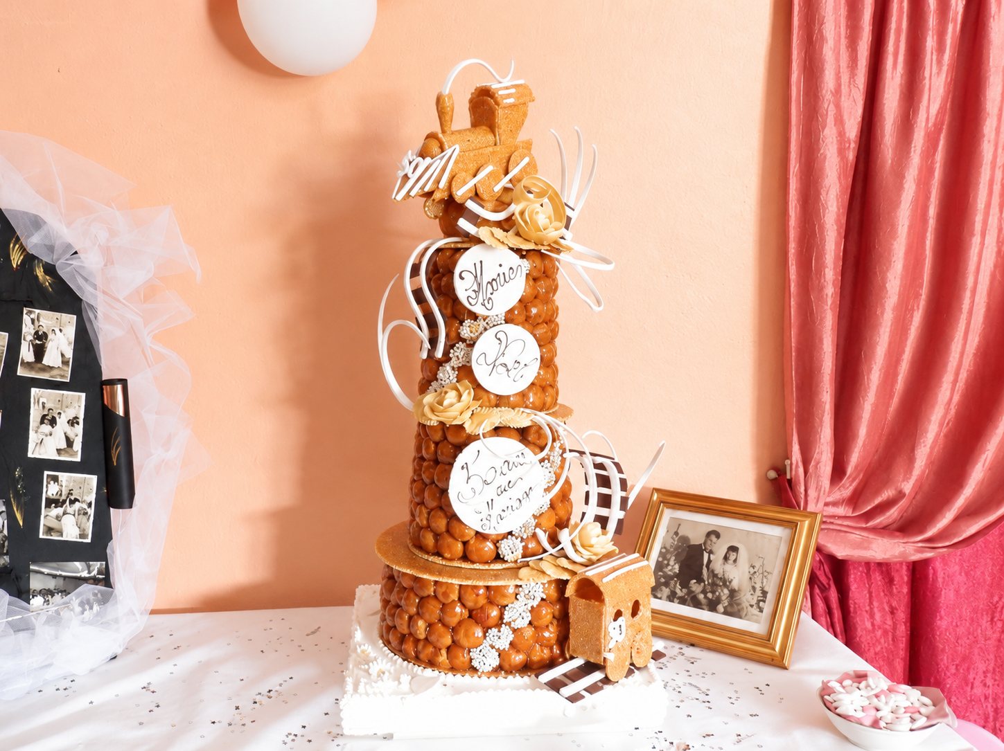 Une grande pièce montée en choux caramélisés ornée de médaillons blancs inscrits à la main, de fleurs en sucre et d'un petit train au sommet, posée sur une table de fête près d'un cadre doré.