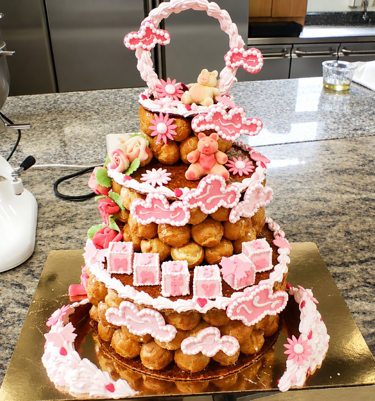 Pièce montée à trois étages de choux caramélisés, ornée de glaçage rose, de fleurs, de petits oursons et de cubes décoratifs, posée sur un plan de travail en granit dans un laboratoire.
