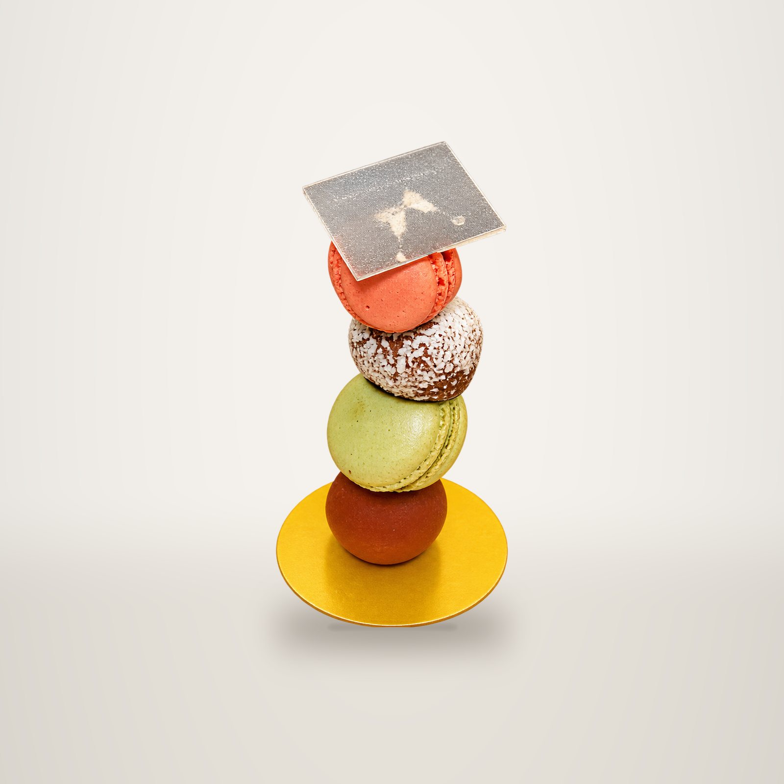 Une pile de macarons de couleurs variées, surmontée d'une petite carte, est posée sur un support jaune devant un fond beige uni.