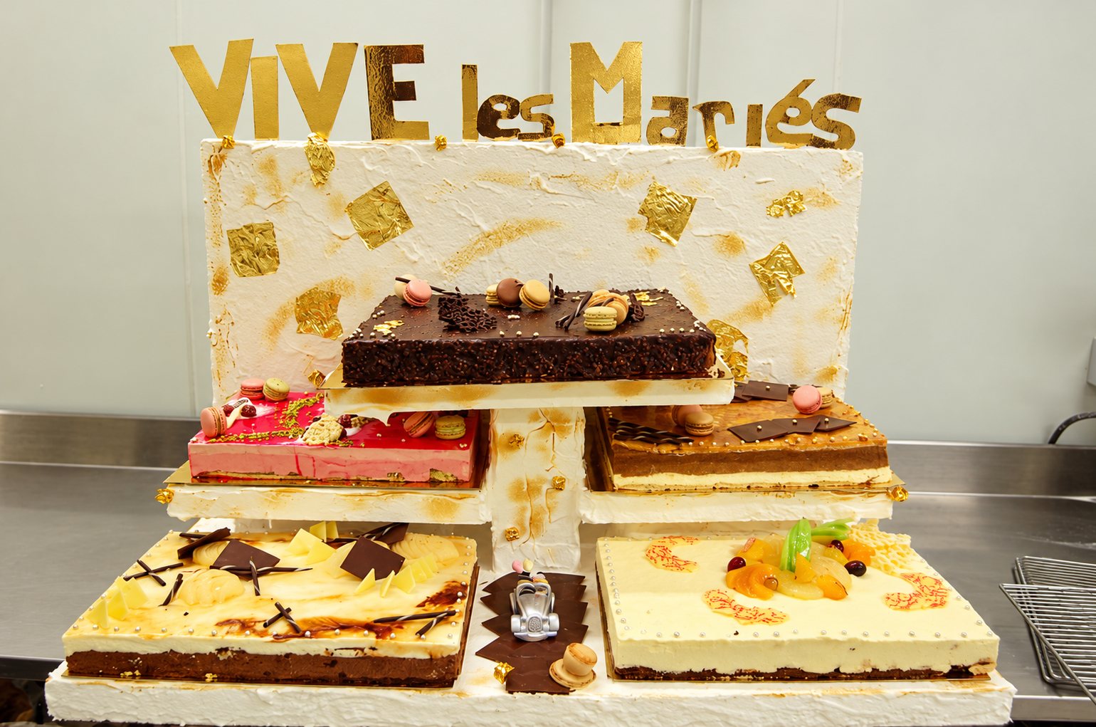 Plusieurs entremets rectangulaires décorés de macarons, chocolat et fruits sont présentés sur un support blanc orné de feuilles d'or, surmonté des lettres dorées « Vive les Mariés ».