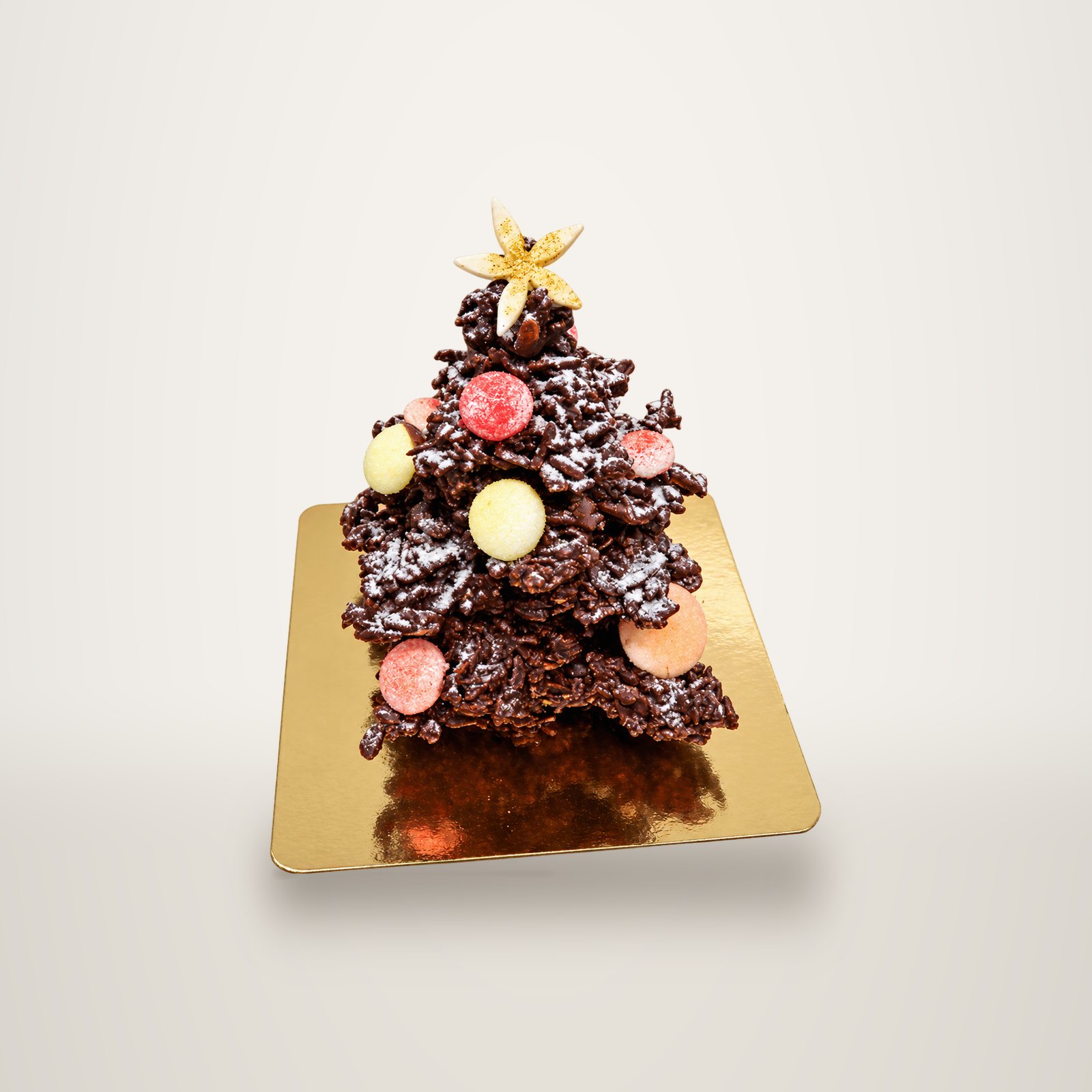 Un petit sapin de Noël en chocolat croustillant saupoudré de sucre glace, décoré de boules colorées et d'une étoile dorée, posé sur un carton doré.