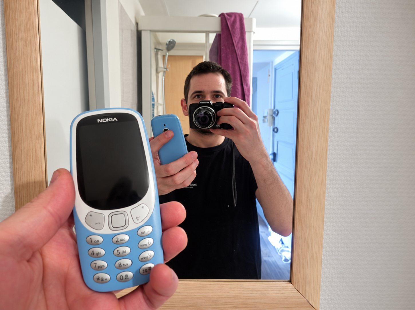 Un homme se photographie dans un miroir avec un appareil photo, tandis qu'une main au premier plan tient un téléphone Nokia bleu à clavier.