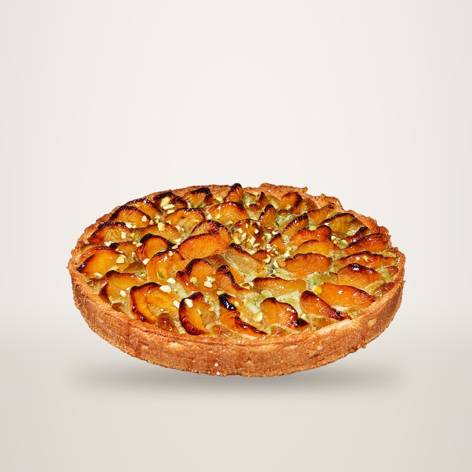 Une tarte à la pâte dorée garnie de moitiés d'abricots sur une crème verte parsemée d'amandes, posée sur un fond bleu uni.